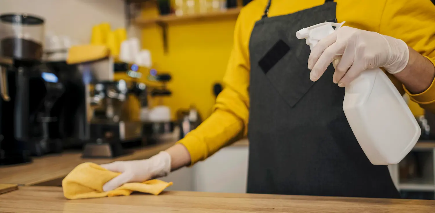 Persona vestida de amarillo y con un delantal negro pasando un trapo con producto desengrasante por una encimera tras aprender a cómo limpiar una cocina industrial”